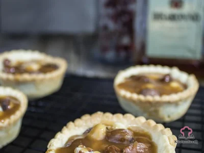 Three almond amaretto tarts on a white marble surface, topped with toasted nuts and glossy amaretto caramel sauce drizzling down the sides