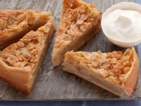 A golden double-crust Almond Apple Pie with steam vents on top, set on a rustic wooden surface with sliced apples and cinnamon sticks nearby
