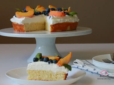 Slice of moist almond cake topped with a thick layer of basil infused whipped cream and garnished with fresh berries and sliced peaches on a white plate