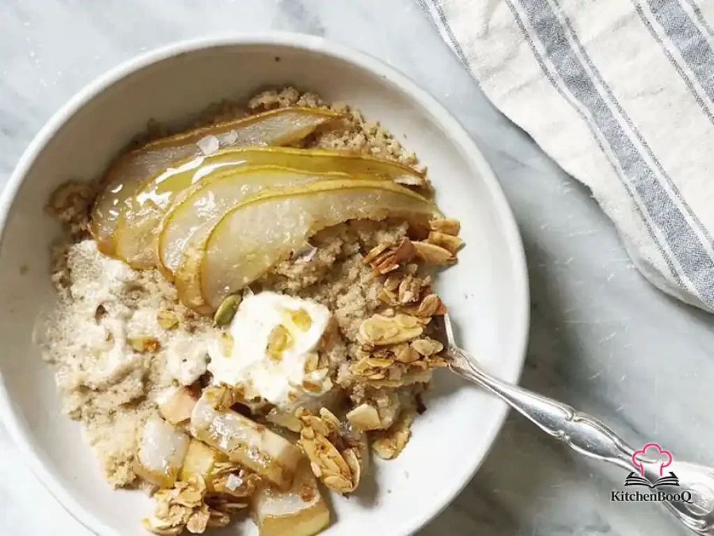 Bowl of creamy amaranth porridge topped with honey-roasted pear slices, a dollop of crème fraîche, crunchy rosemary granola, and a drizzle of olive oil on a rustic wooden surface