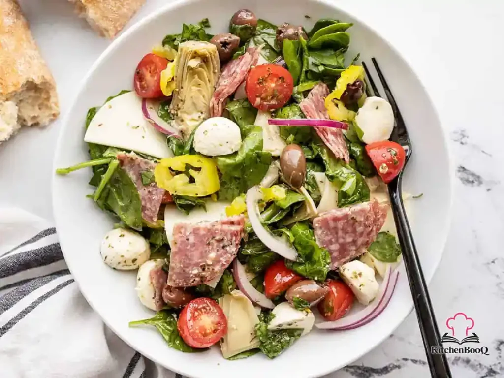 A large bowl of antipasto salad with salami, mozzarella, provolone, artichoke hearts, kalamata olives, grape tomatoes, banana peppers, and red onion on a bed of mixed greens drizzled with homemade Italian dressing