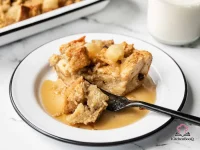 Golden baked apple cinnamon bread pudding in a white casserole dish topped with dollops of applesauce and a dusting of cinnamon