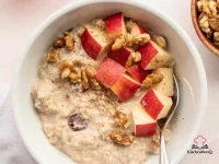 Four glass jars of apple pie overnight oats topped with cinnamon, chopped walnuts, and dried cranberries on a wooden board