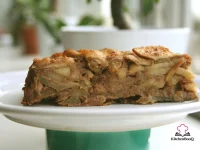 Rustic golden Apple and Amaretti Cake on a white cake stand, showing a golden-brown top crust with visible apple slices and amaretti crumble