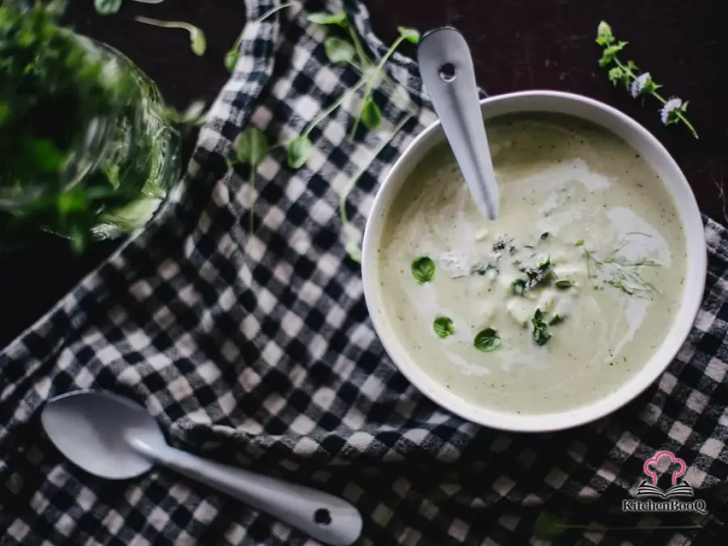 A white bowl of smooth creamy apple and fennel soup garnished with chopped mint, fennel fronds, and a swirl of cream on a rustic wooden table