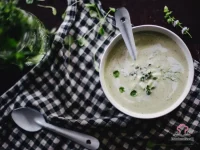 A white bowl of smooth creamy apple and fennel soup garnished with chopped mint, fennel fronds, and a swirl of cream on a rustic wooden table