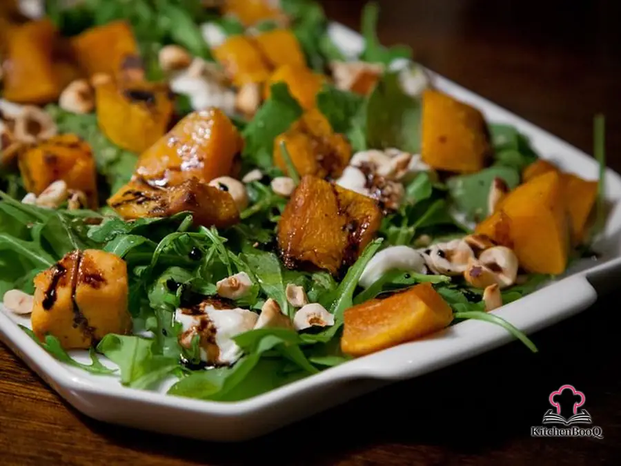 Platter of arugula salad topped with roasted butternut squash cubes, sliced burrata, toasted halved hazelnuts, and a drizzle of dark balsamic glaze on a rustic wooden board