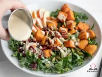 A large bowl of autumn kale and sweet potato salad topped with roasted sweet potato cubes, apple slices, pepitas, chopped pecans, dried cranberries, and drizzled with creamy maple tahini dressing