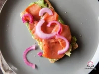 Two pieces of rye crispbread topped with smashed avocado, slices of lox, and vibrant quick-pickled red onions on a white plate