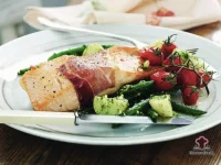 Prosciutto-wrapped baked salmon fillet served with basil pesto chat potatoes, green beans, and roasted cherry tomatoes on a white plate