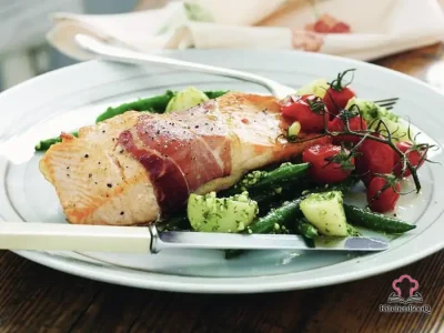Prosciutto-wrapped baked salmon fillet served with basil pesto chat potatoes, green beans, and roasted cherry tomatoes on a white plate