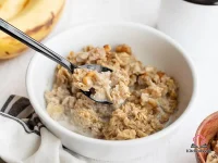 Slice of banana bread baked oatmeal in a white casserole dish topped with sliced bananas and walnuts on a wooden table
