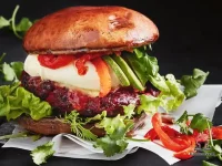 A stack of bean and beetroot mushroom burgers topped with melted mozzarella, sliced avocado, fresh coriander, and sour cream on a white plate