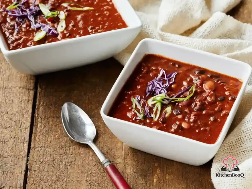 A rustic bowl of beef and black bean chili topped with sliced green onions served alongside toasted baguette slices on a wooden table