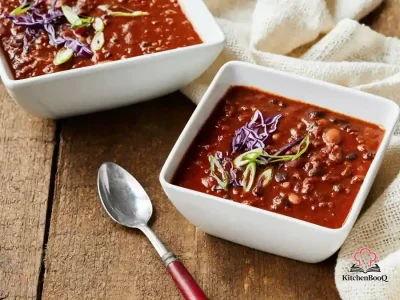 A rustic bowl of beef and black bean chili topped with sliced green onions served alongside toasted baguette slices on a wooden table