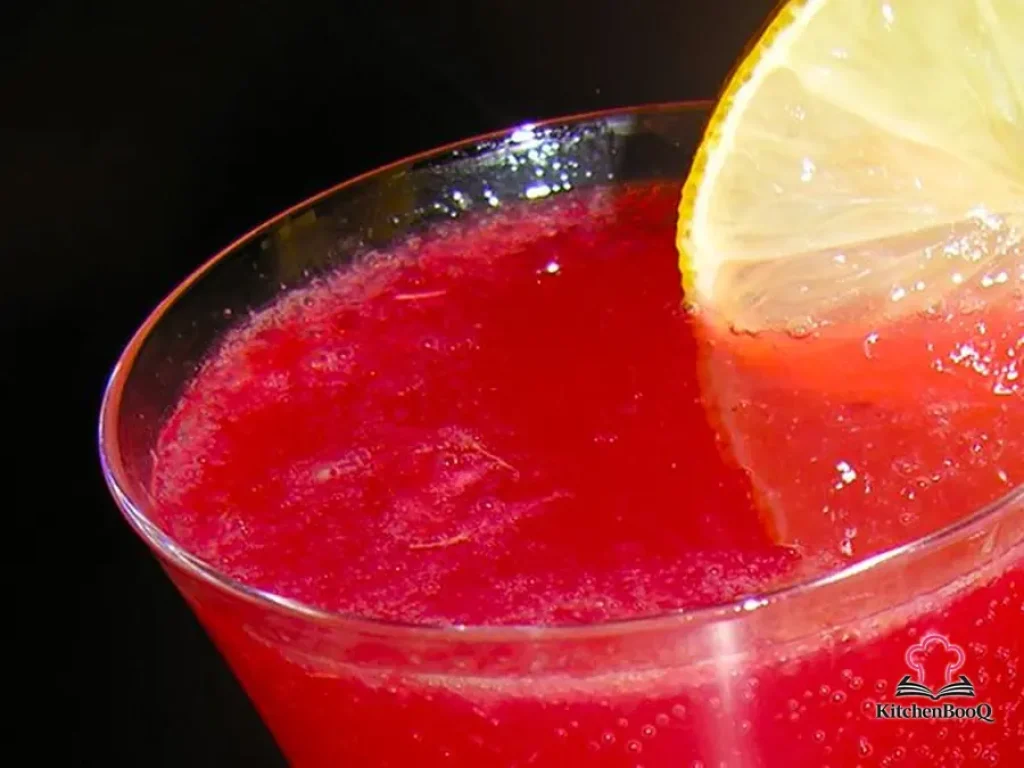 A Prosecco flute filled with deep ruby Bloody Ginger cocktail garnished with a Meyer lemon slice on a white marble surface