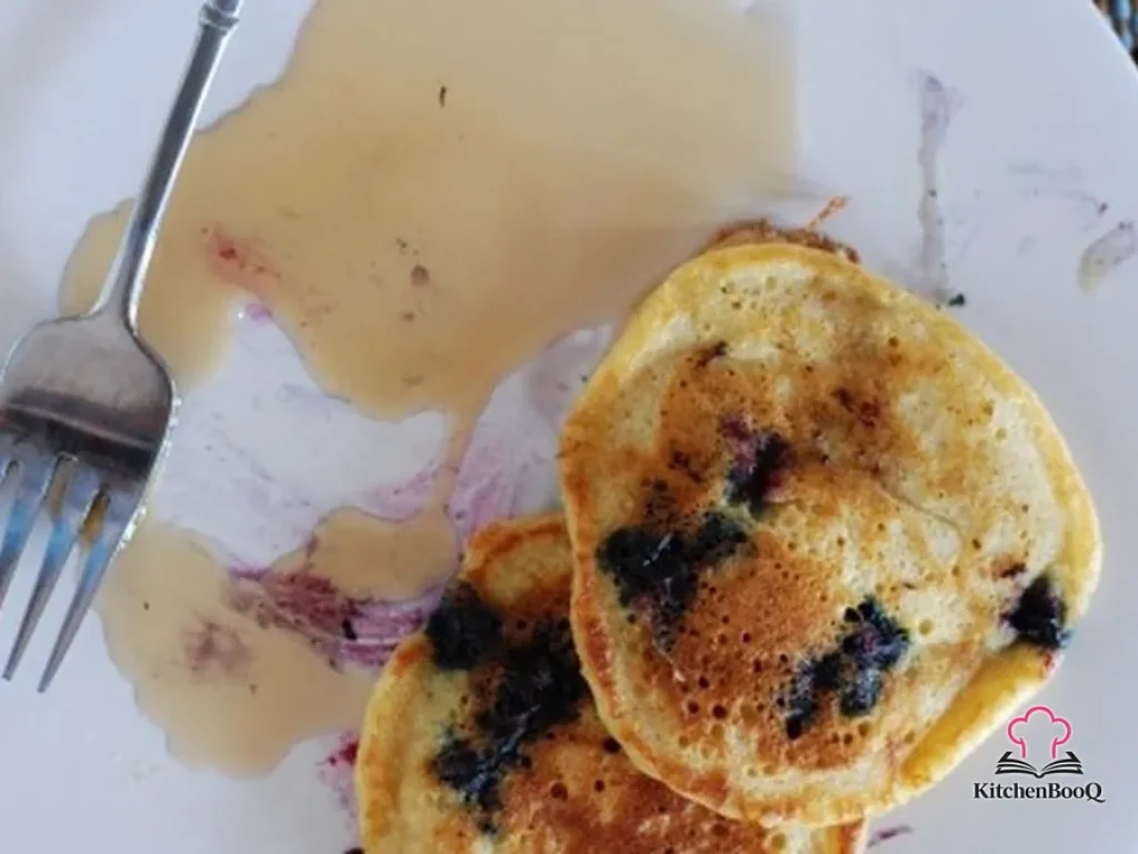 Stack of fluffy blueberry buttermilk hotcakes on a white plate drizzled with maple syrup and dusted with powdered sugar, with fresh blueberries scattered around