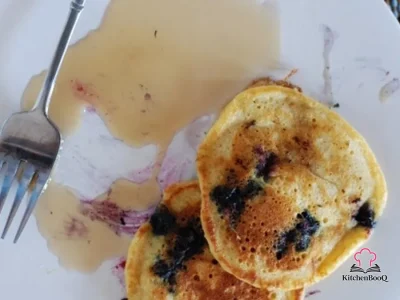 Stack of fluffy blueberry buttermilk hotcakes on a white plate drizzled with maple syrup and dusted with powdered sugar, with fresh blueberries scattered around