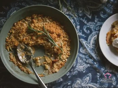Golden bourbon rosemary apple crisp in a white oval baking dish with bubbling caramel apple filling and crunchy oat crumble topping, served with a dollop of whipped cream