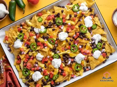 Overhead view of a baking sheet loaded with breakfast nachos topped with scrambled eggs, melted cheddar cheese, black beans, diced tomato, sliced jalapeño, red onion, and dollops of sour cream