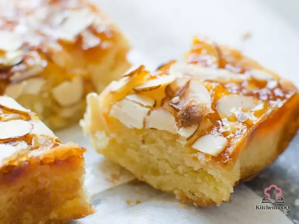 Apricot almond bars cut into squares on a wooden cutting board, showing a golden almond paste base topped with glossy apricot preserves and toasted sliced almonds