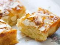 Apricot almond bars cut into squares on a wooden cutting board, showing a golden almond paste base topped with glossy apricot preserves and toasted sliced almonds