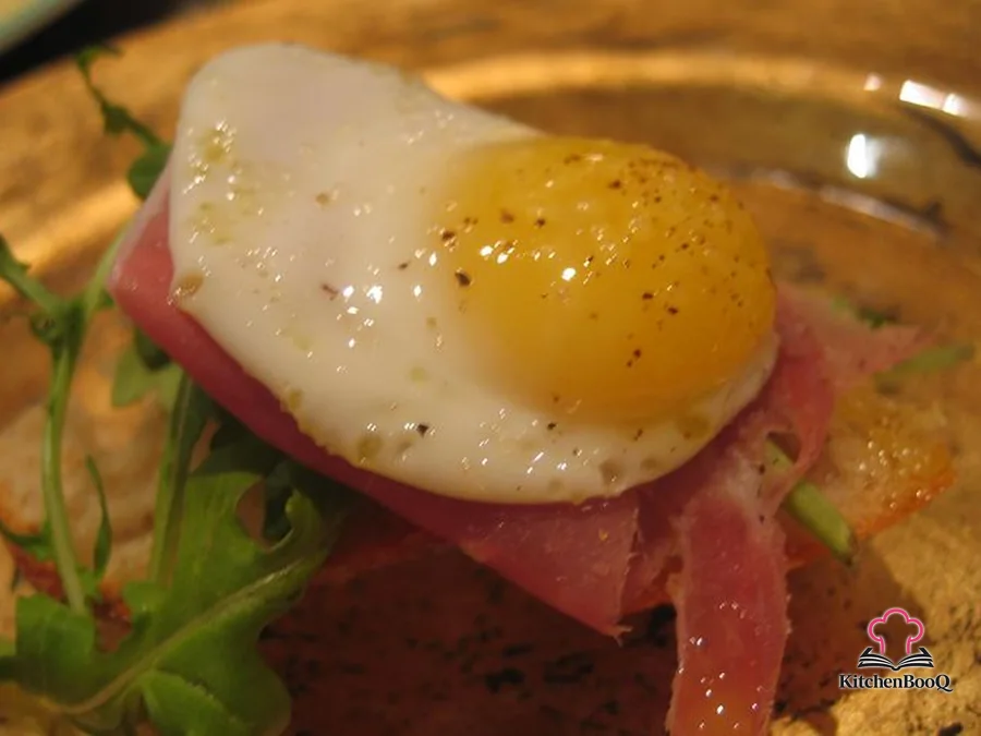 Six baguette rounds topped with folded prosciutto, baby arugula, and fried quail eggs sprinkled with pink sea salt and cracked pepper on a rustic wooden board