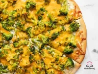 Broccoli cheddar pizza with herby white sauce cut into eight slices on a round pizza pan with melted golden cheddar and bright green broccoli florets