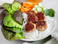 A bowl of Bún Chả with golden caramelized pork meatballs, rice vermicelli noodles, fresh butter lettuce, mint, sliced carrots, cucumber, and a small bowl of nuoc cham dipping sauce on the side