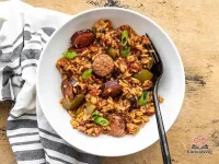 A cast iron skillet filled with Cajun sausage and rice, garnished with sliced green onions, showing golden browned Andouille sausage slices nestled in seasoned red rice
