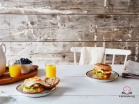 A bánh mì breakfast sandwich on a toasted English muffin stacked with roasted chicken thigh, fried egg, sliced avocado, julienned cucumber, pickled daikon and carrot do chua, and fresh cilantro leaves, served on a wooden board