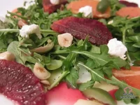 A composed salad of baby arugula topped with blood orange and Cara Cara orange segments, green Castelvetrano olives, crumbled goat cheese, and chopped toasted hazelnuts, drizzled with olive oil on a white dinner plate