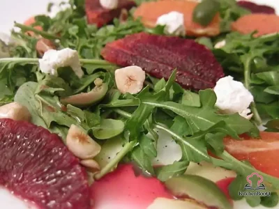 A composed salad of baby arugula topped with blood orange and Cara Cara orange segments, green Castelvetrano olives, crumbled goat cheese, and chopped toasted hazelnuts, drizzled with olive oil on a white dinner plate
