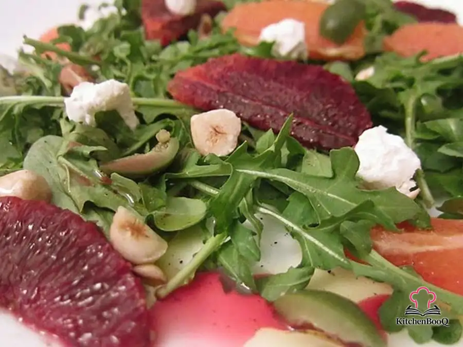 A composed salad of baby arugula topped with blood orange and Cara Cara orange segments, green Castelvetrano olives, crumbled goat cheese, and chopped toasted hazelnuts, drizzled with olive oil on a white dinner plate