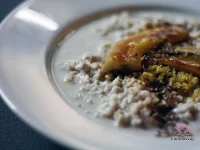 A bowl of creamy coconut milk oatmeal topped with caramelized banana halves, flax seeds, lemon zest, and a drizzle of maple syrup on a rustic wooden surface