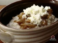 A white bowl of creamy steel cut oats topped with golden caramelized onions and a dollop of fresh white ricotta cheese on a rustic wooden surface