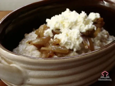 A white bowl of creamy steel cut oats topped with golden caramelized onions and a dollop of fresh white ricotta cheese on a rustic wooden surface