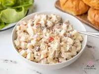 A bowl of creamy chicken salad with diced apple, dried cranberries, and red onion, served on a white plate with a fork