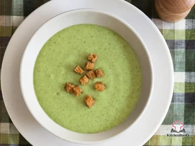 A bowl of creamy cream of broccoli soup garnished with small broccoli florets and a swirl of cream on a rustic wooden table