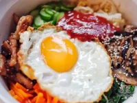 A large bowl of bibimbap with white rice topped with sautéed carrots, wilted spinach, gochujang cucumber, sliced beef, and a sunny-side-up egg drizzled with sesame oil and sesame seeds