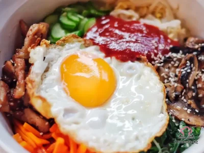 A large bowl of bibimbap with white rice topped with sautéed carrots, wilted spinach, gochujang cucumber, sliced beef, and a sunny-side-up egg drizzled with sesame oil and sesame seeds