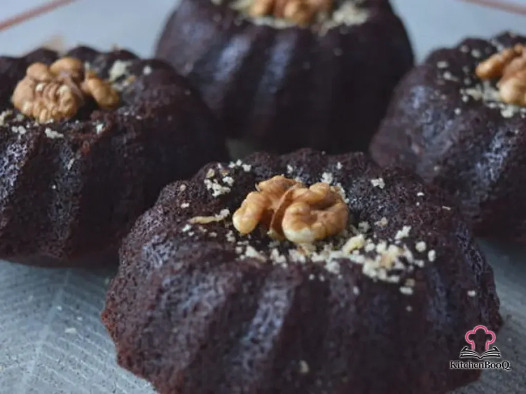 Four dark chocolate mini cakes dusted with chopped walnuts on a rustic wooden board, showing a dense moist crumb
