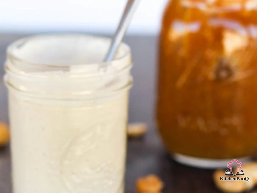 A small glass jar filled with creamy cashew and date coffee creamer next to a cup of black coffee and scattered cashews and medjool dates on a wooden surface