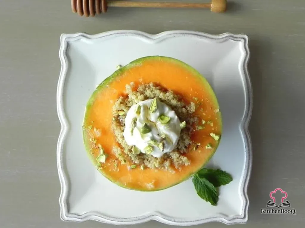 Charentais melon half stuffed with cardamom-spiced quinoa topped with Greek yogurt, crushed pistachios, fresh mint leaves, and a honey drizzle on a white plate