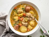 A large pot of hearty chicken stew with tender chicken thighs, baby potatoes, sliced carrots, and celery in a thick golden-brown broth, garnished with fresh chopped parsley