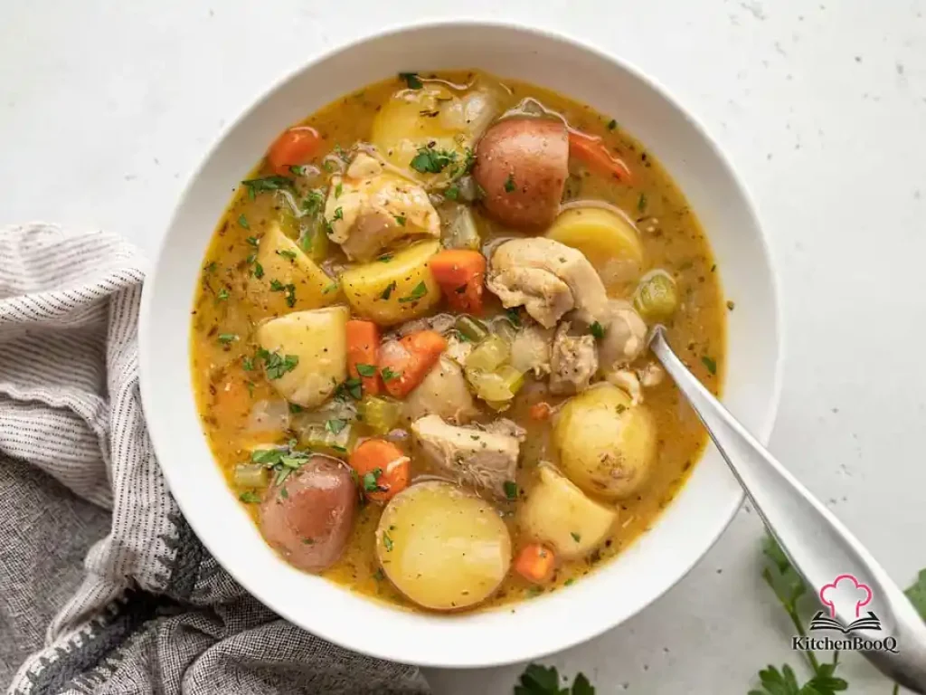 A large pot of hearty chicken stew with tender chicken thighs, baby potatoes, sliced carrots, and celery in a thick golden-brown broth, garnished with fresh chopped parsley