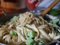 Bowl of whole grain fettuccine coated in creamy chickpea Alfredo sauce topped with asparagus, spinach, and peas garnished with fresh basil and shaved Parmesan