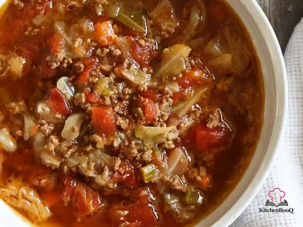 A rustic bowl of chunky beef cabbage and tomato soup with visible pieces of ground beef, green cabbage, carrots, and celery in a rich red broth