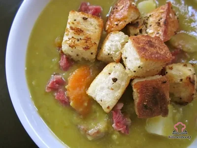 A bowl of chunky split pea soup with shredded ham, diced carrots, and potato pieces in a thick green broth, served with crusty bread on a rustic wooden table