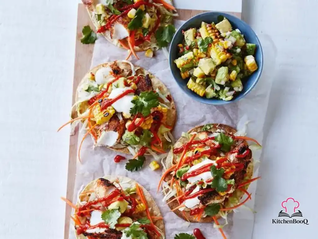 Crispy corn tortilla tostadas topped with flaky Mexican-seasoned flathead fish, charred corn and avocado salsa, shredded iceberg lettuce, and carrot matchsticks served on a white plate with lime wedges